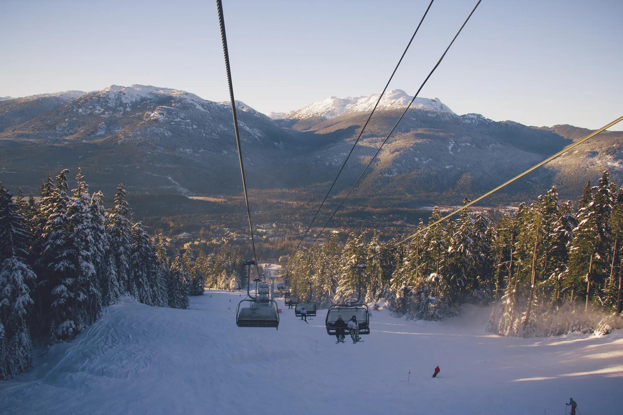 Skiers riding a chairlift up a snowy slope, with mountains behind