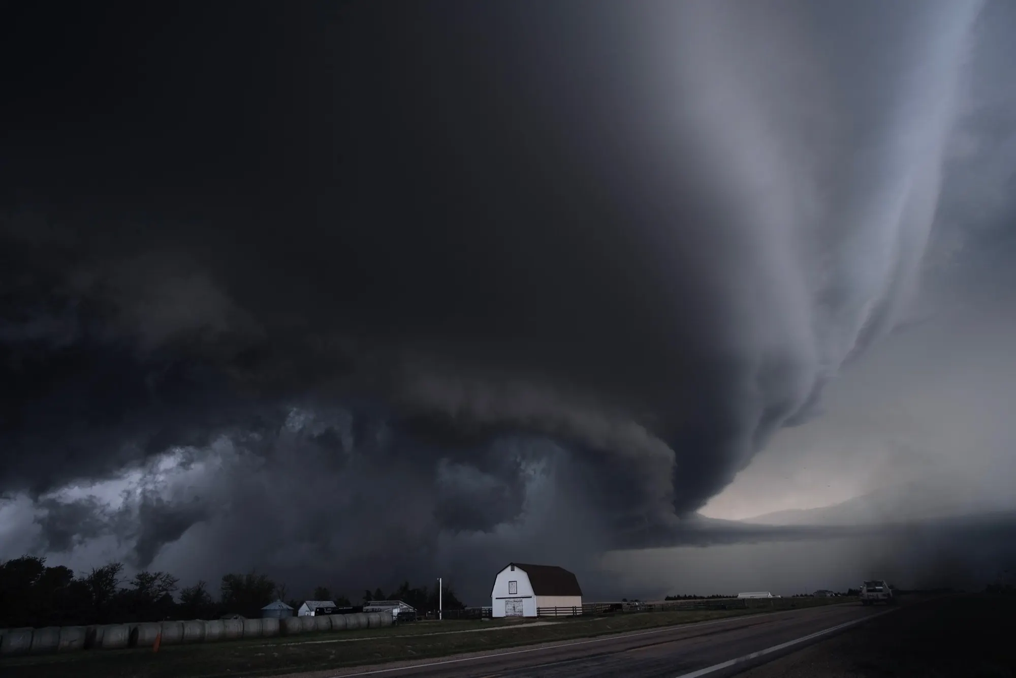 A supercell thunderstorm over a farm road and a white barn in Kansas